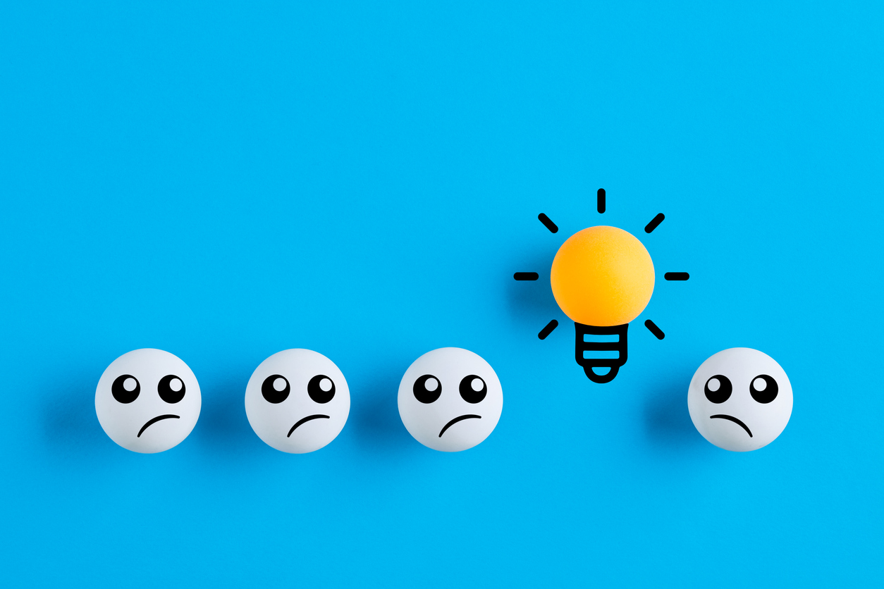 A row of white ping pong balls with concerned faces drawn on. a yellow ping pong ball shaped like a light bulb stands out from the rest of the group.